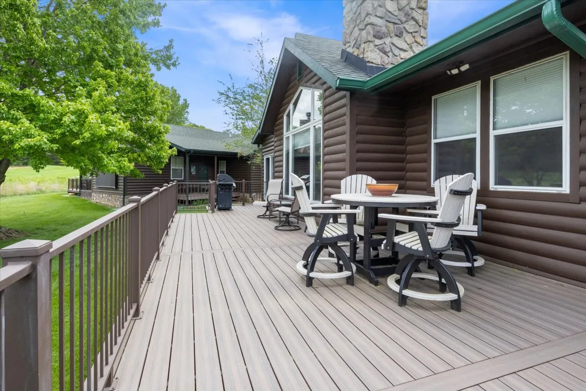 This image showcases a spacious deck area featuring comfortable seating, a gas grill, and a view of a landscaped yard against a rustic log cabin facade. The space is set up for outdoor dining and socializing, with plenty of room for families or groups of friends to gather. The grill is positioned for easy access, and the deck overlooks green space, ideal for relaxing or watching activity on the grounds beyond.