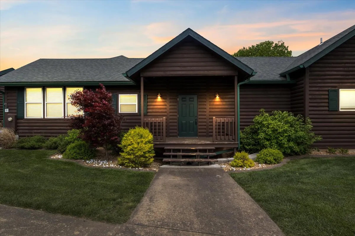 This exterior shot captures the inviting front door of a log cabin-style home, set amidst a landscaped garden. The porch area is perfect for morning coffees or evening relaxation, and the log texture evokes a sense of rustic luxury consistent with a golf resort.