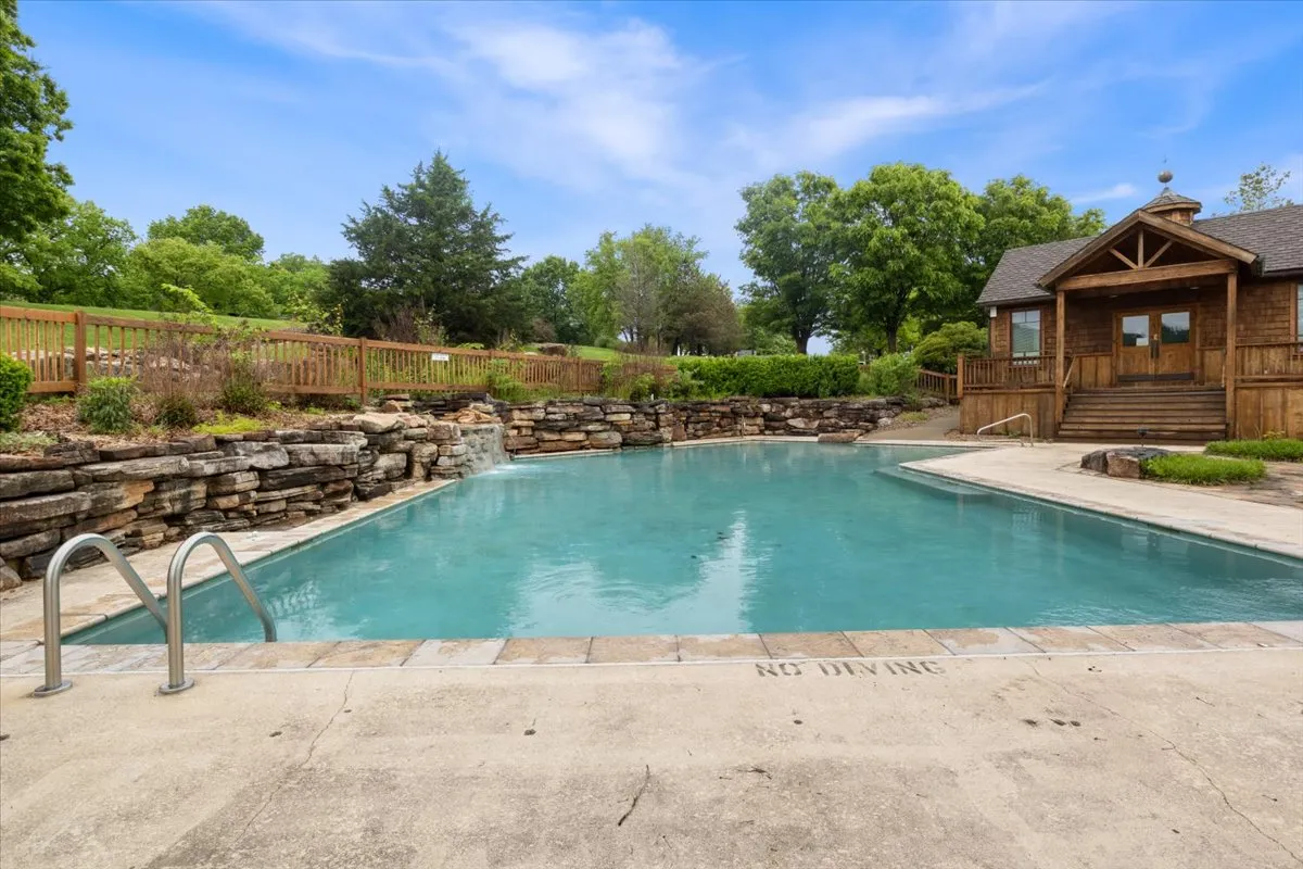 A vibrant pool area featuring a wooden cabin, a stone wall, and abundant surrounding greenery. The scene is lively and relaxed, perfect for swimming, sunbathing, or group barbecues.