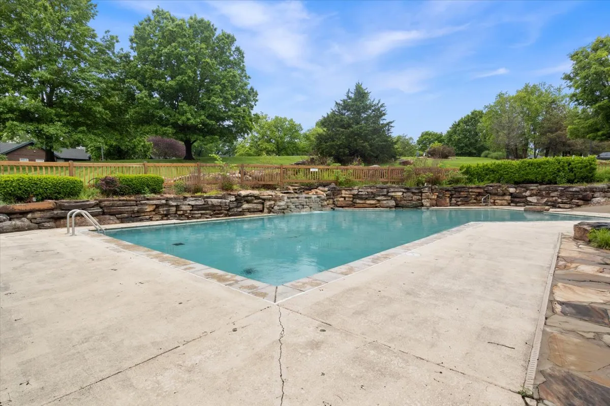 A sparkling community swimming pool framed by a stone wall and surrounded by lush greenery under a clear blue sky. The pool area is spacious, inviting, and designed for families to gather, swim, sunbathe, and enjoy outdoor activities together.