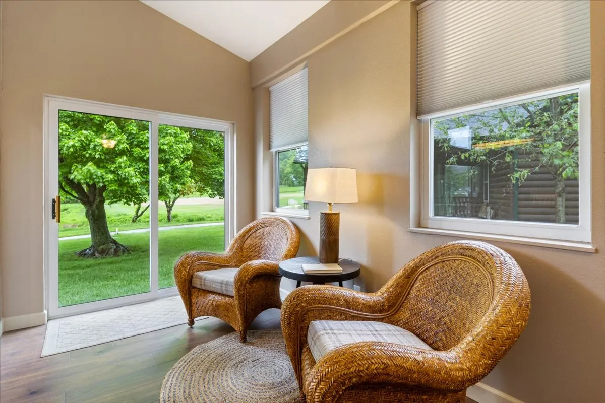 The image features a pair of wicker chairs situated by a table lamp, set in front of large windows that offer expansive views of the outdoors. The natural light streaming in emphasizes the connection to the surrounding landscape, while the seating arrangement suggests a cozy spot to relax and watch wildlife or changing seasons.