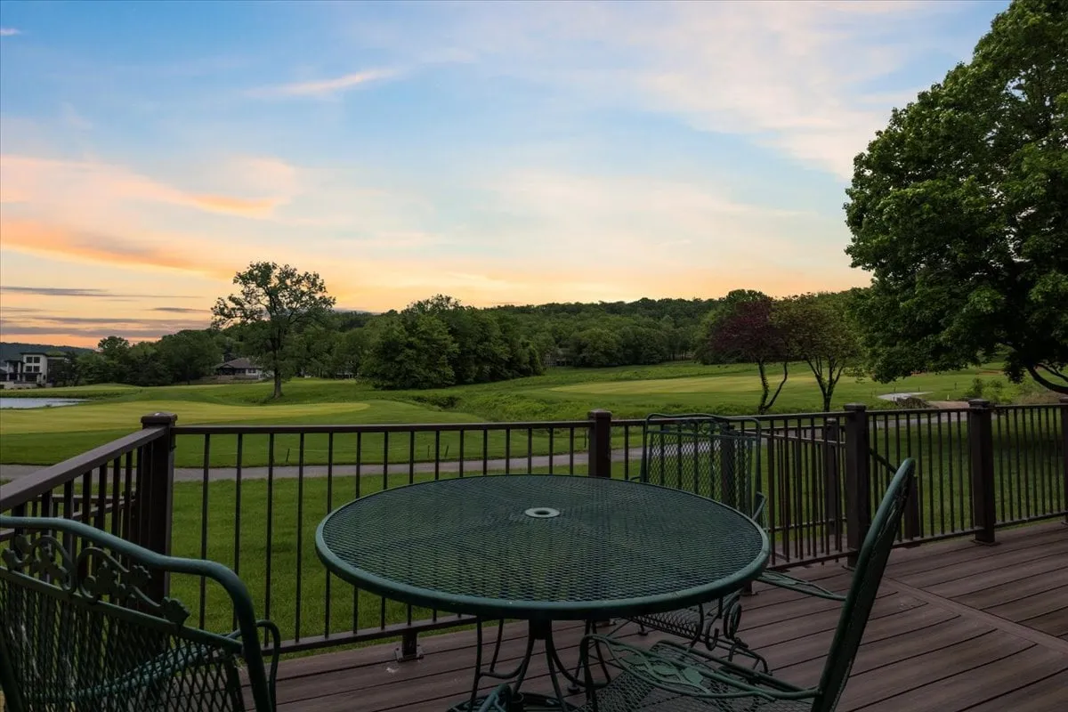 The image showcases an inviting outdoor dining area with a table and chairs set on a patio, surrounded by lush greenery and distant trees. The scene is bathed in the warm glow of a sunset sky, creating a tranquil atmosphere perfect for relaxation or gathering with friends and family. The expansive lawn and serene landscape highlight the cottage's peaceful golf course setting.