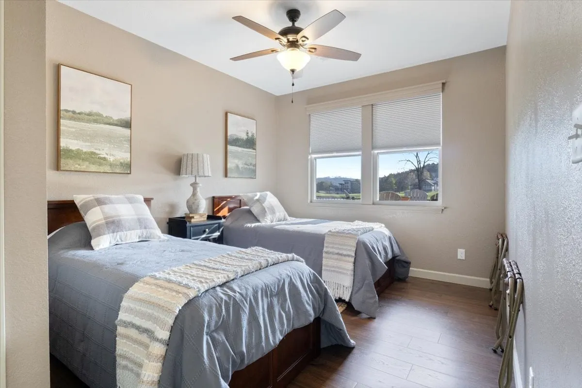This interior bedroom scene features two twin beds dressed in soft bedding, a window providing plenty of natural light and a view outside, a decorative lamp, and tranquil wall art. The overall mood is relaxed and inviting, with a setup ideal for kids, friends, or teammates sharing the space.