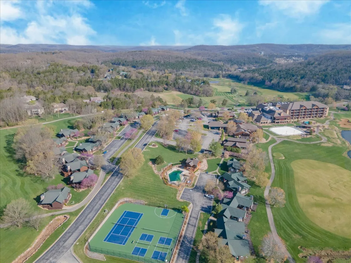 This exterior image captures the community's tennis courts and swimming pool set amidst lush greenery, with wooden cabins and a hotel building nearby. The scene is lively yet serene, suggesting abundant recreational activities and the inviting atmosphere of a resort community.