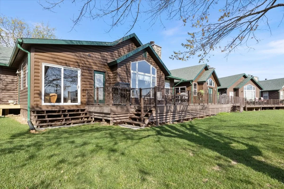 The image displays a classic log cabin structure with expansive windows and a spacious deck area, ideal for gatherings and relaxing. The setting highlights outdoor living and blends seamlessly with the natural environment of a golf resort.