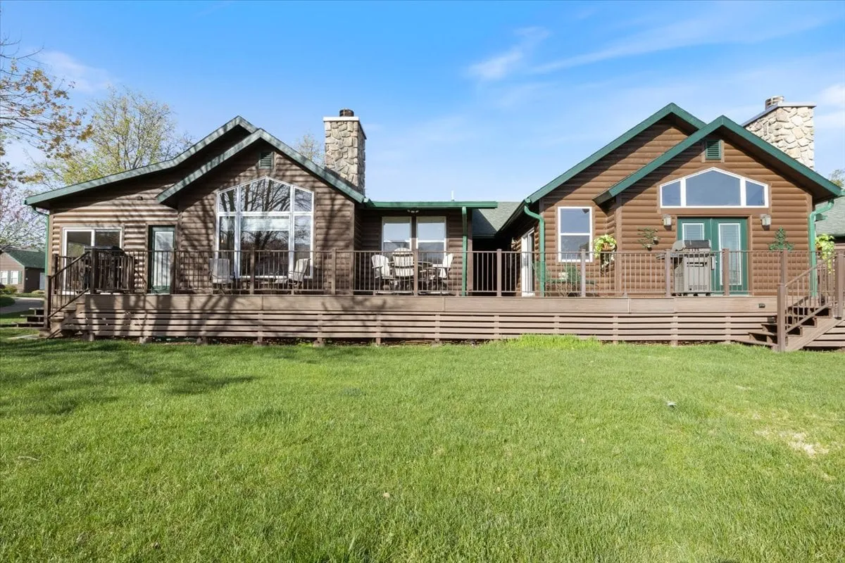 This exterior view focuses on large windows and a stone chimney, with the deck area visible as a place for outdoor gatherings. The mood is inviting, capturing the golden light of sunset and suggesting moments of relaxation as the day winds down. The deck is clearly a transition space between the comfort of the interior and the beauty of the outdoors.
