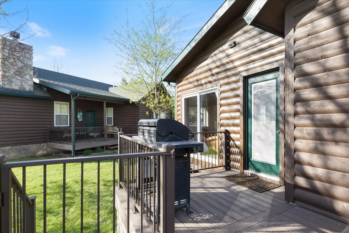 The photo features the deck of a log cabin, with a grill ready for barbecuing, a green lawn for play or relaxation, and the inviting architecture of the cottage itself. The scene is cozy but open, suggesting both family fun and tranquil privacy. The deck connects fluidly to the outdoors, supporting activities like alfresco dining or evening stargazing.